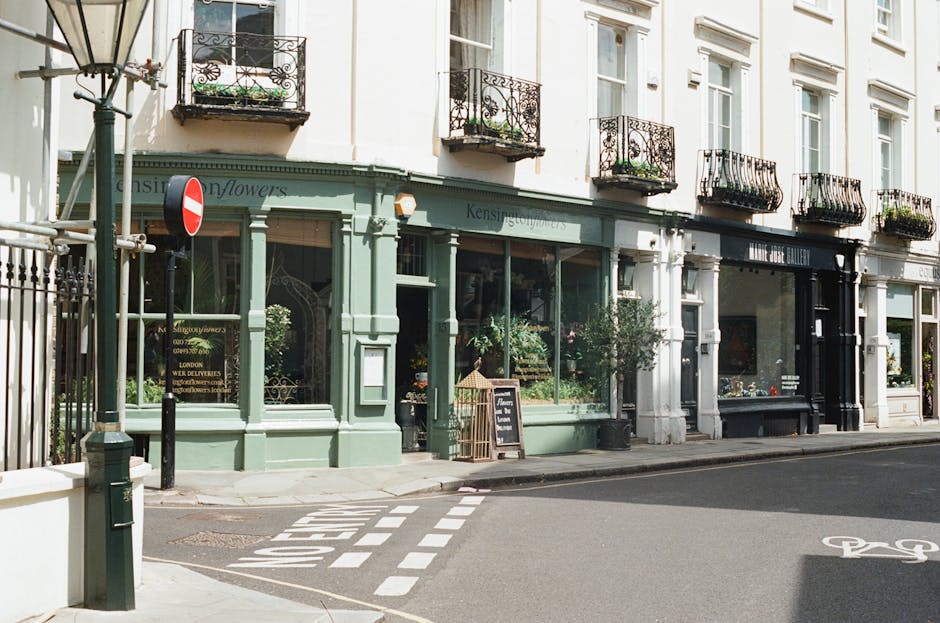 A floral shop with a light green façade, featuring large front windows displaying growing plants and flower arrangements inside. The shop has decorative black wrought-iron balconies on the upper floo